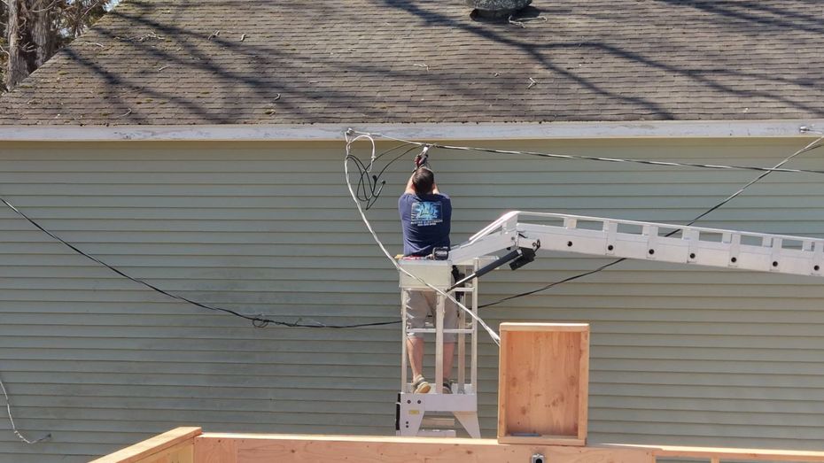 Worker standing on a lift beside a building, reaching toward overhead power lines.