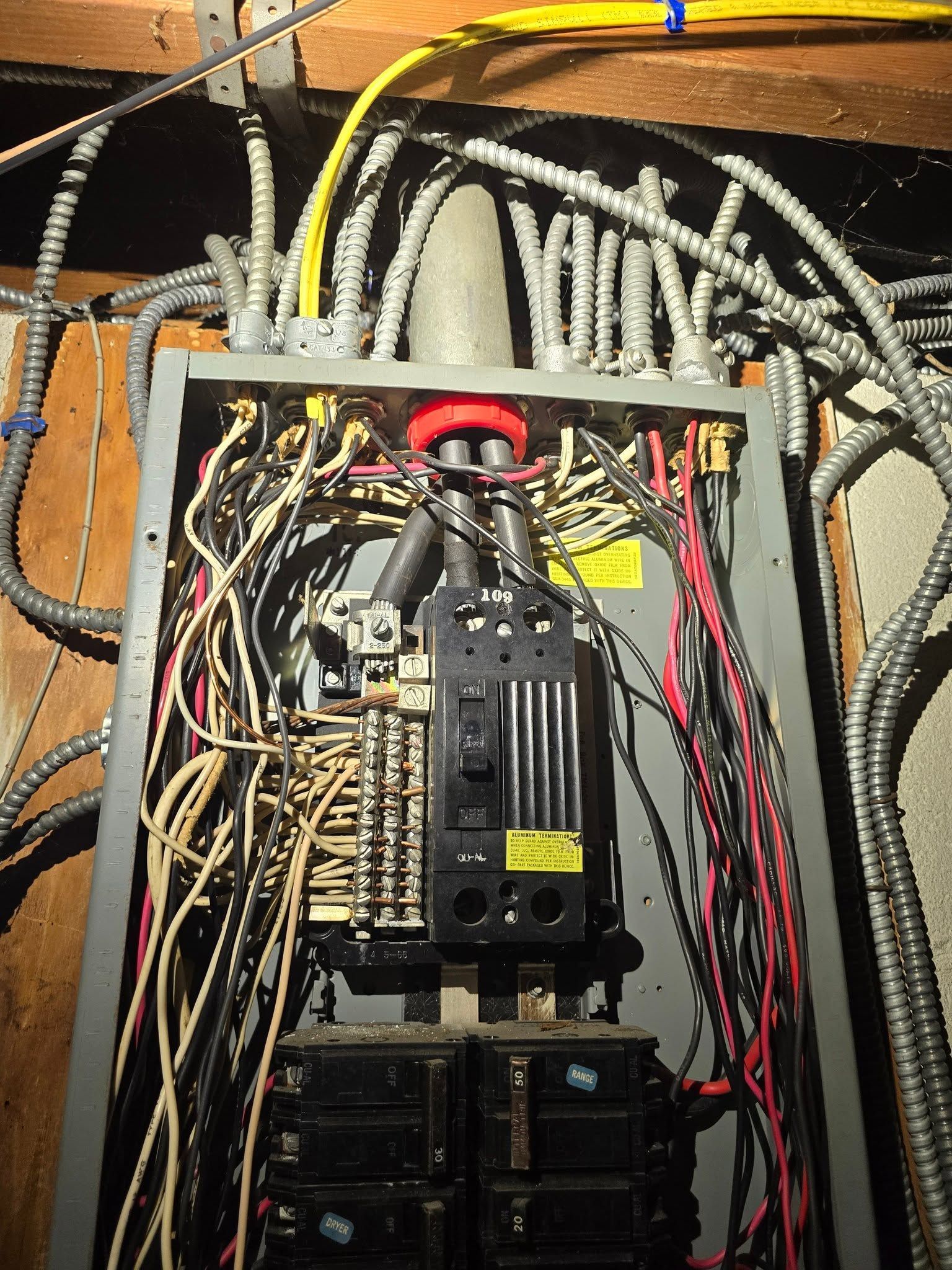 Electrical panel with dense gray wiring and circuit breakers inside a wooden cabinet, with a red cable loop.