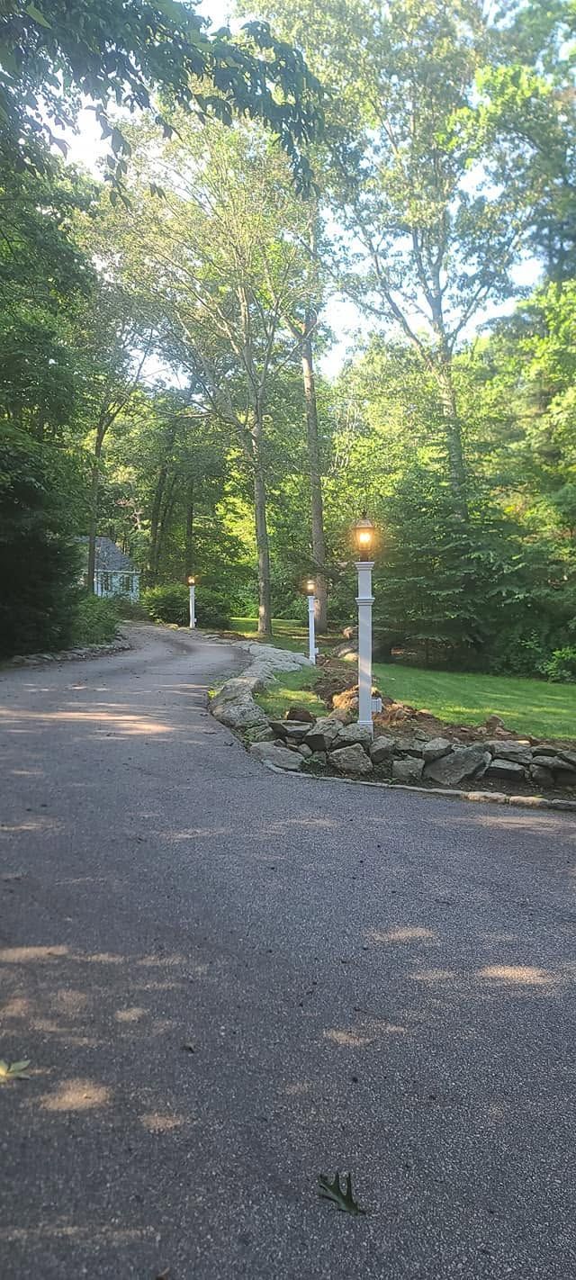 Gravel driveway curving through a wooded garden, with a stone border and lit lamppost at dusk