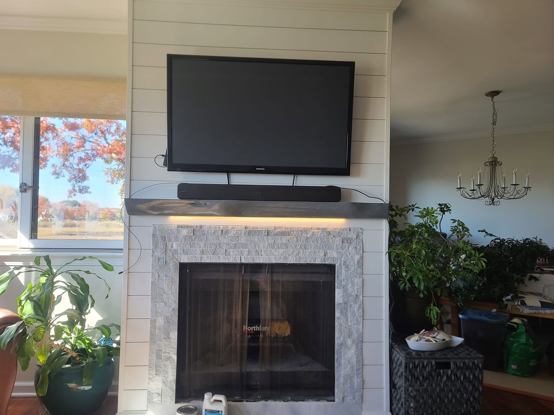 Living room fireplace with a wall-mounted TV, white brick surround, and houseplants near bright windows