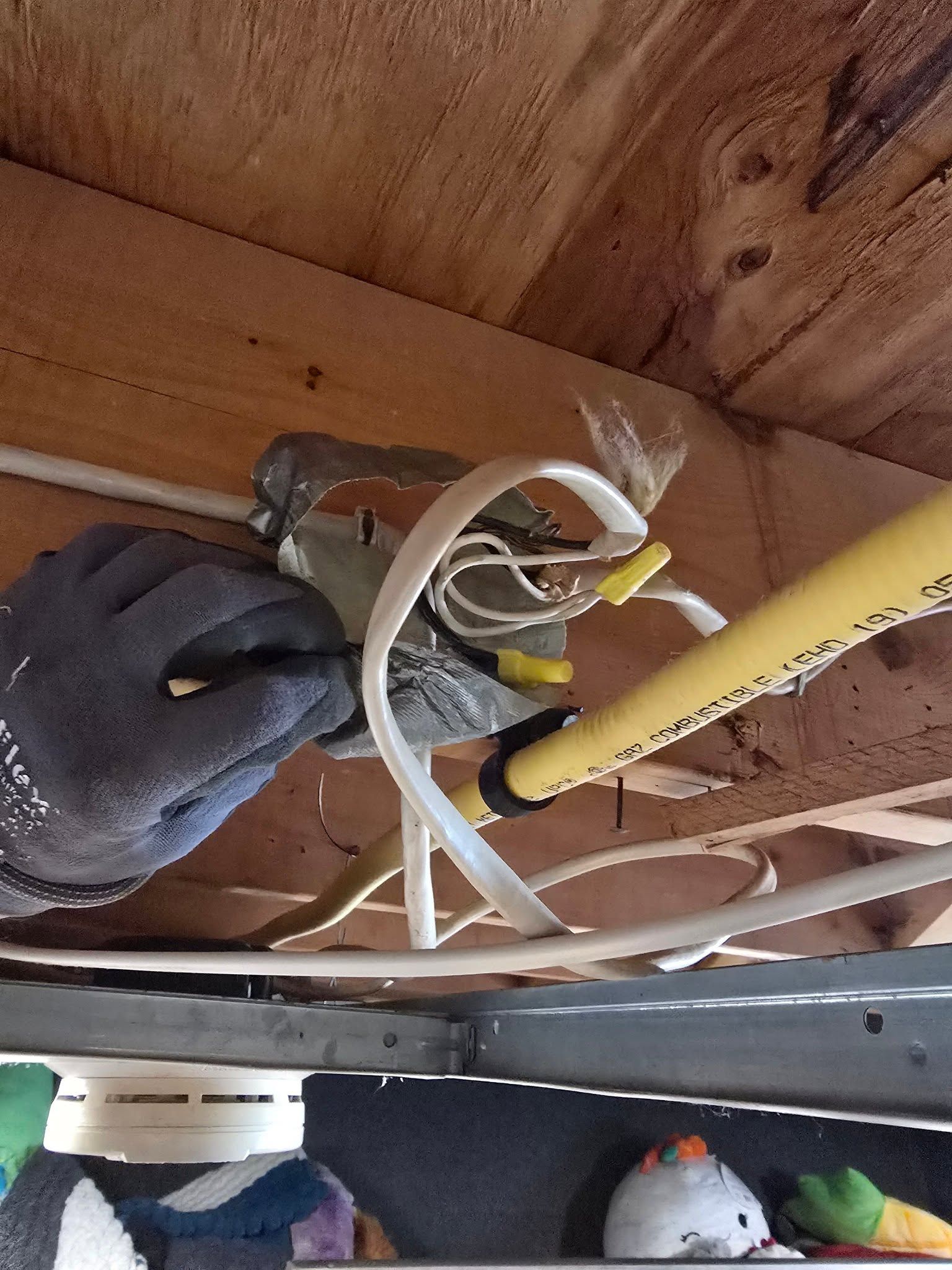Gloved hand working on electrical wiring inside a wooden ceiling cavity with yellow and white cables