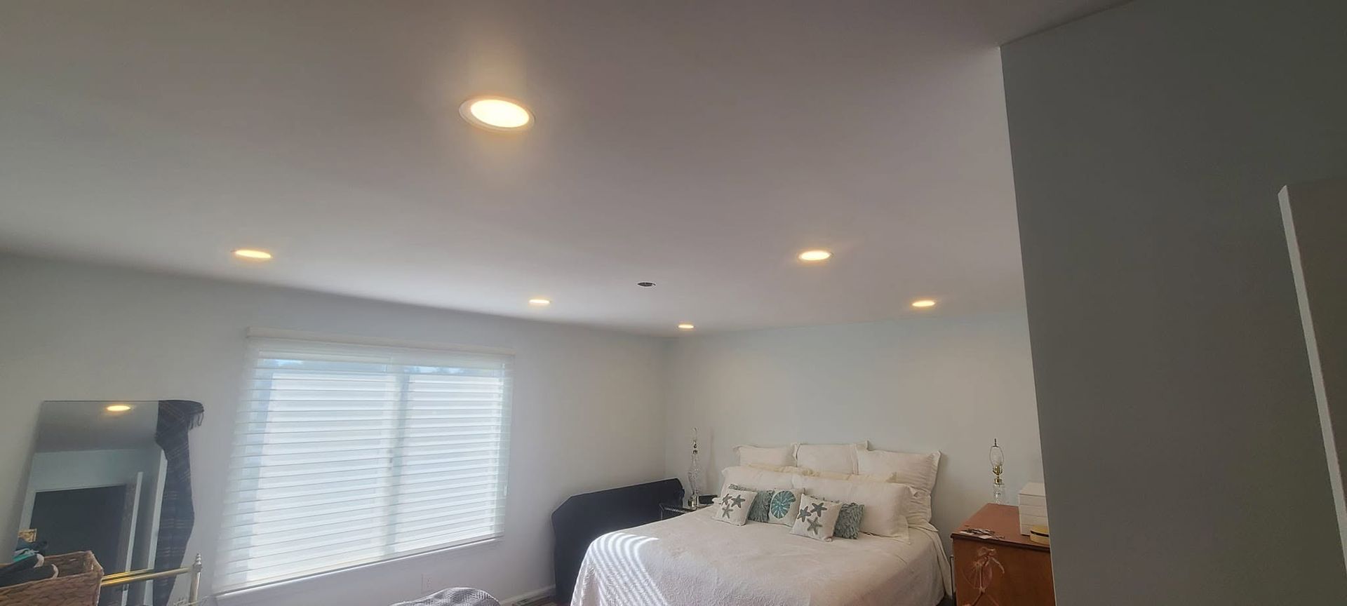 Bright bedroom with a bed, white blinds, recessed lights, and a dresser near the window.