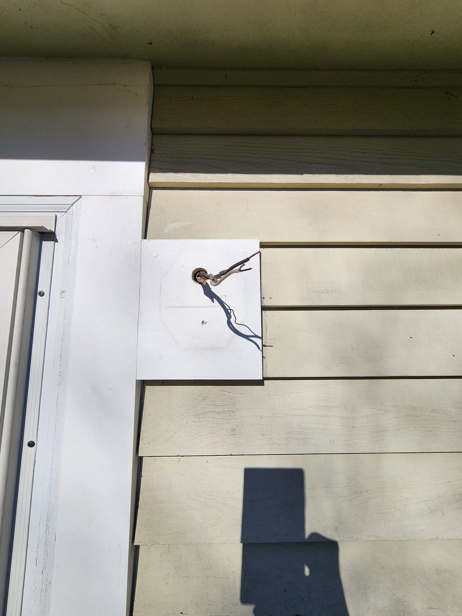White sign with a small metal fixture mounted on beige house siding near a door.