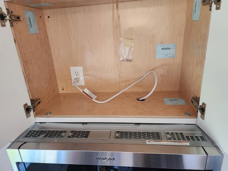 Open wooden cabinet with a white cord plugged into an outlet above a stainless appliance.