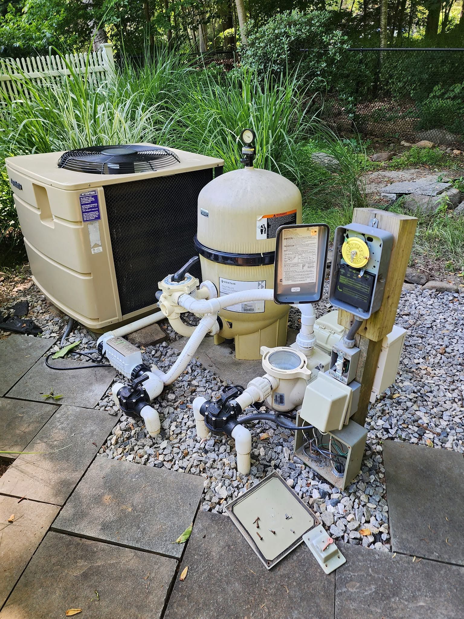 Outdoor pool equipment and plumbing on a patio beside a beige unit, with greenery in the background.