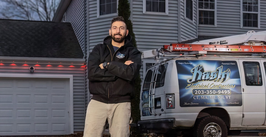 Man standing with arms crossed beside a service van and garage, with a ladder on the van roof.