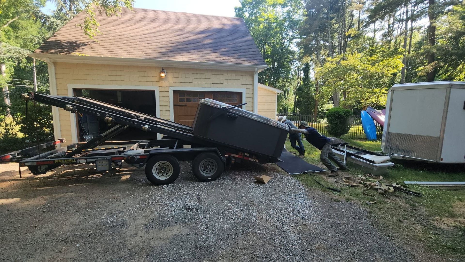 Black dump trailer tipped in a driveway beside a garage, with trees and a small shed nearby.