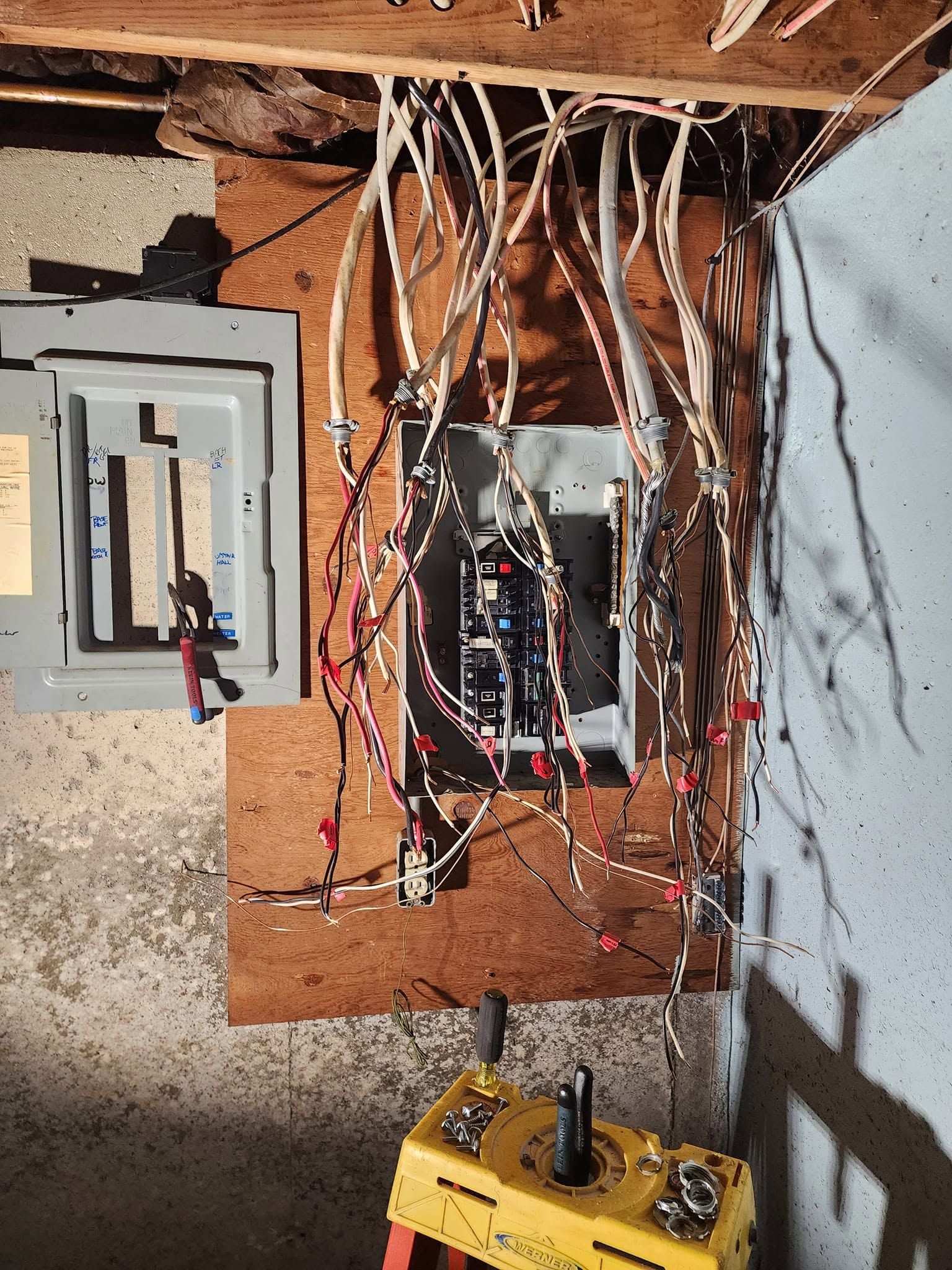 Exposed wall wiring and electrical panels in an unfinished room, with a yellow meter on the floor.
