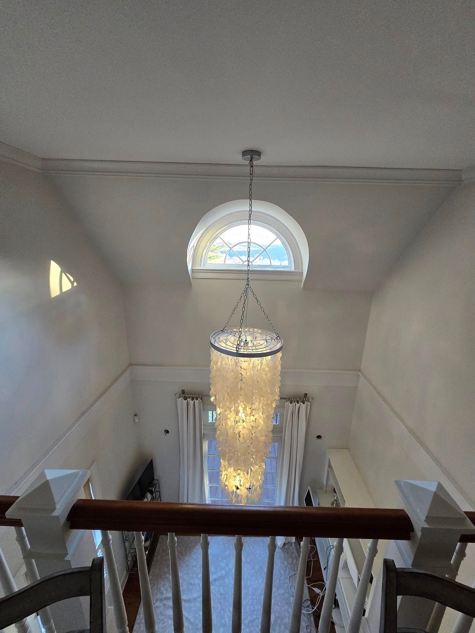 Staircase landing with a hanging chandelier and arched window, viewed from above through a white railing.
