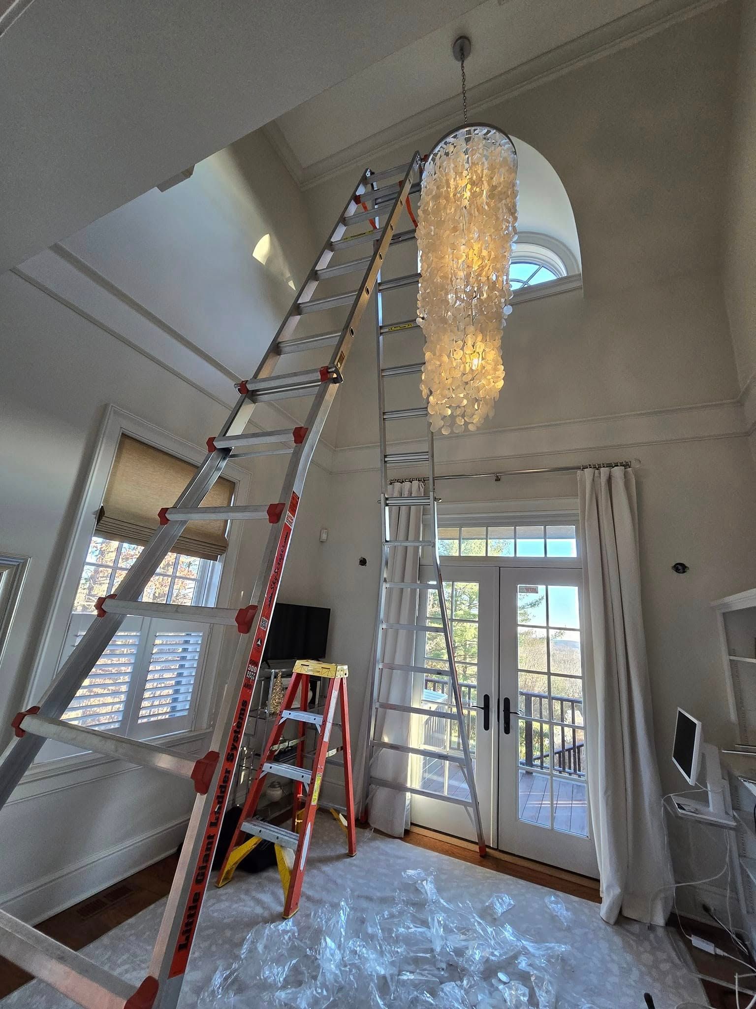 Tall ladder setup in a bright entryway with a hanging chandelier and a smaller ladder below; work in progress.
