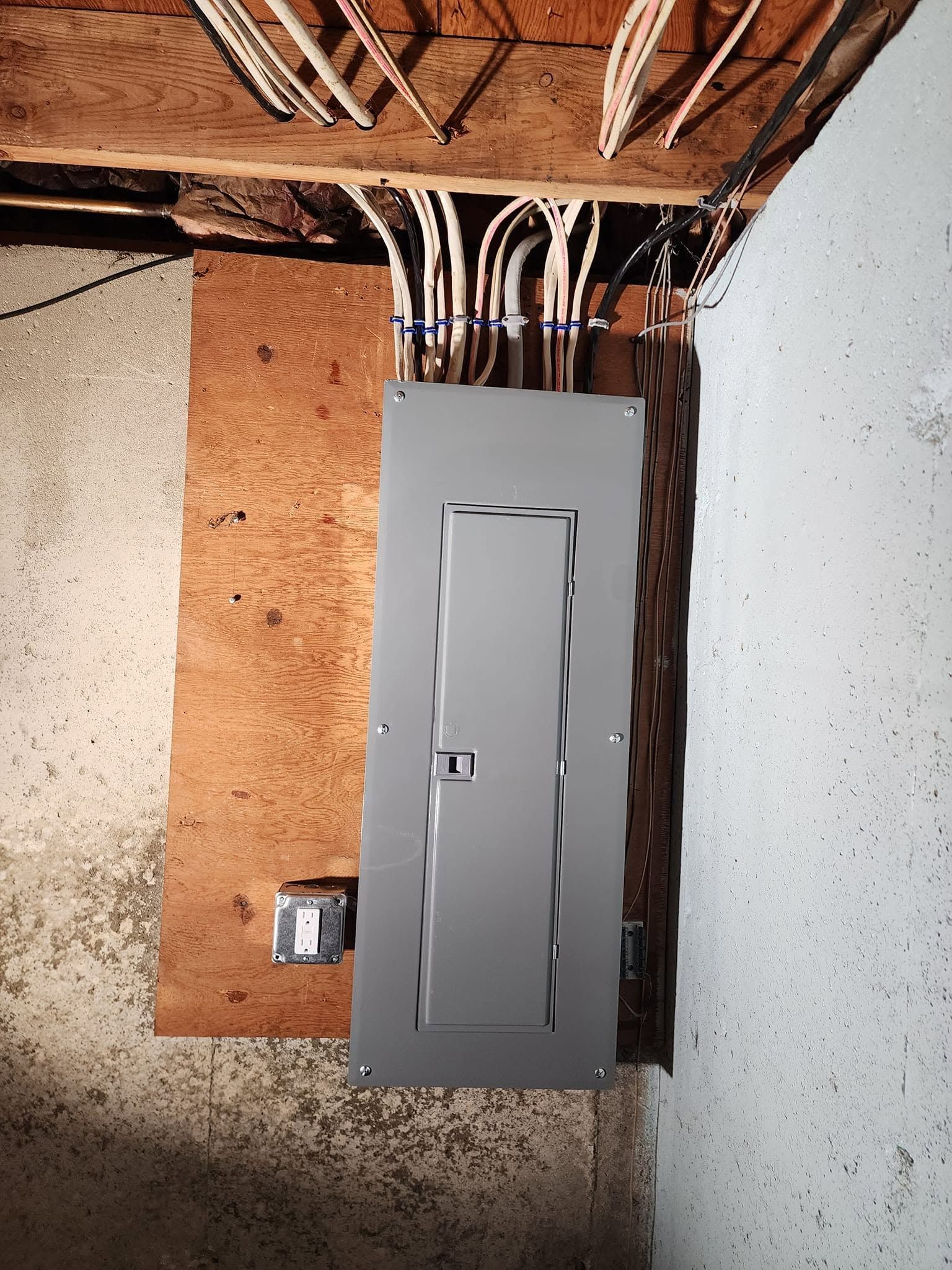 Electrical breaker panel mounted on a plywood board with exposed wiring above it.