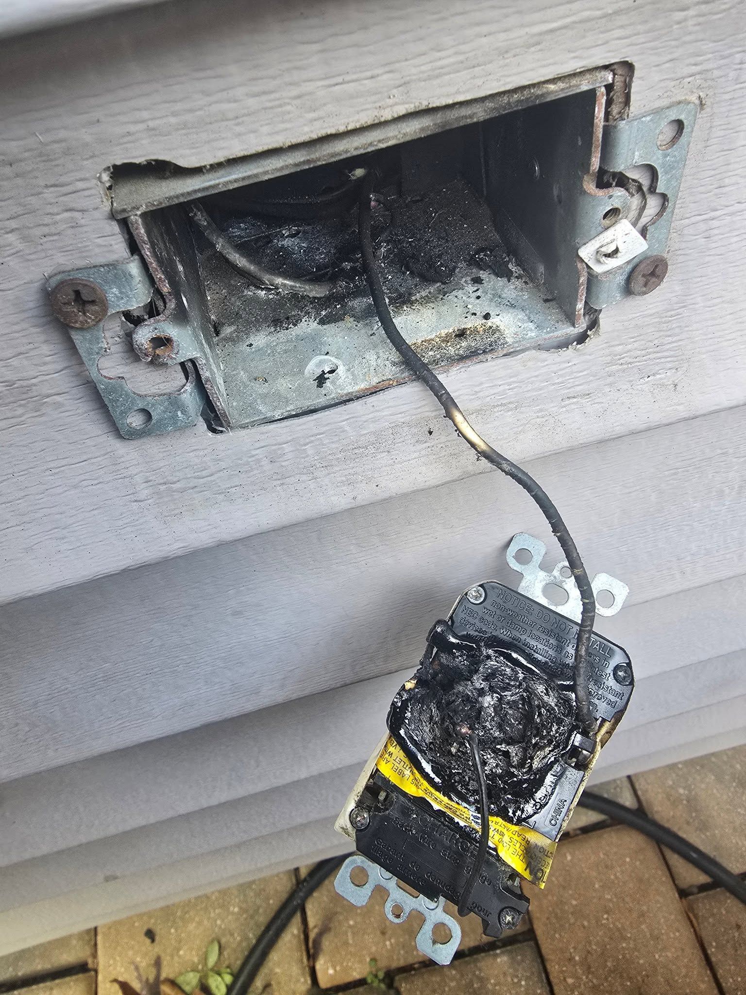Burned electrical outlet box with a damaged black device hanging by wires near a wall.