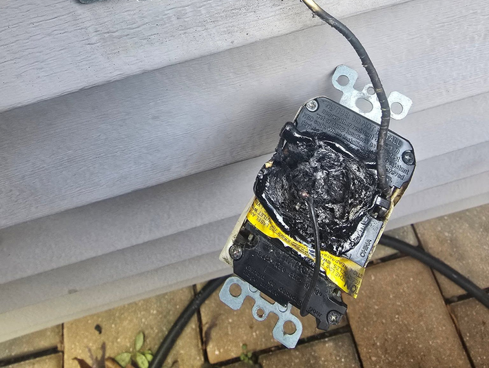 Black electrical outlet with yellow tape hanging off a gray wall siding outdoors