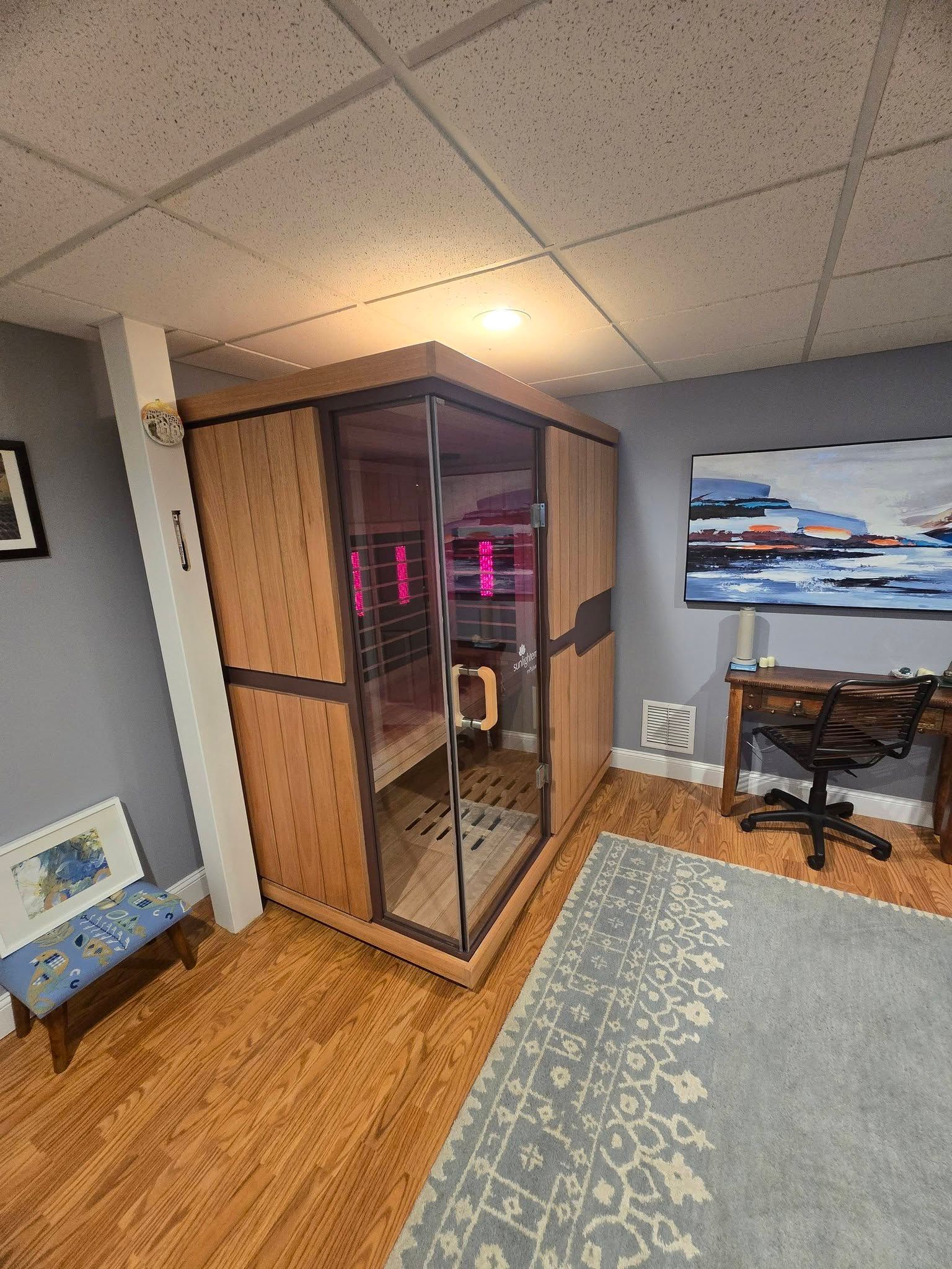 Small office with a glass booth, desk chair, wall art, and hardwood floor in a basement-like room.
