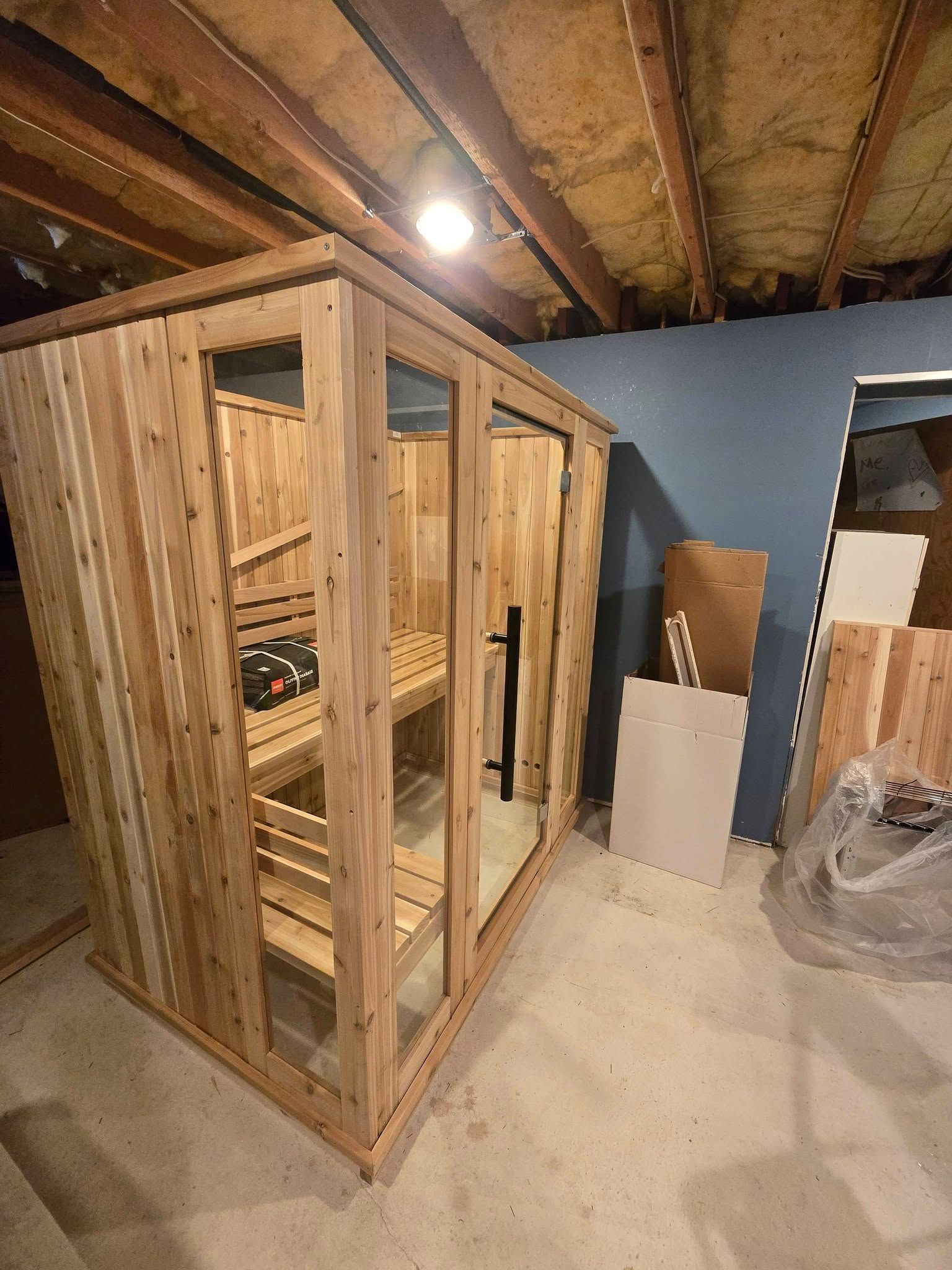 Wooden sauna room under construction in a basement, with framed walls, glass door, and insulation visible.