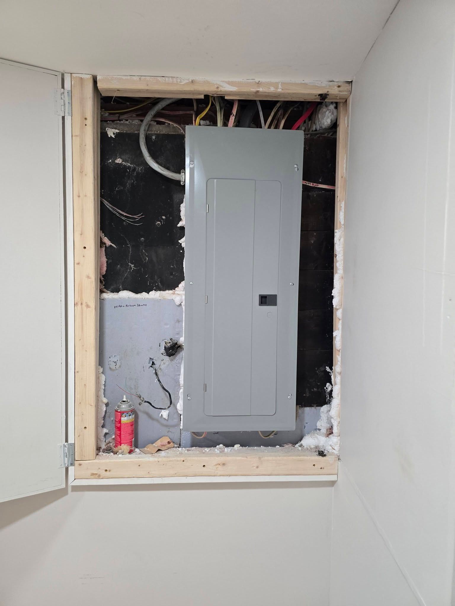 Electrical panel in unfinished wall opening with exposed wiring and drywall, partially painted interior.