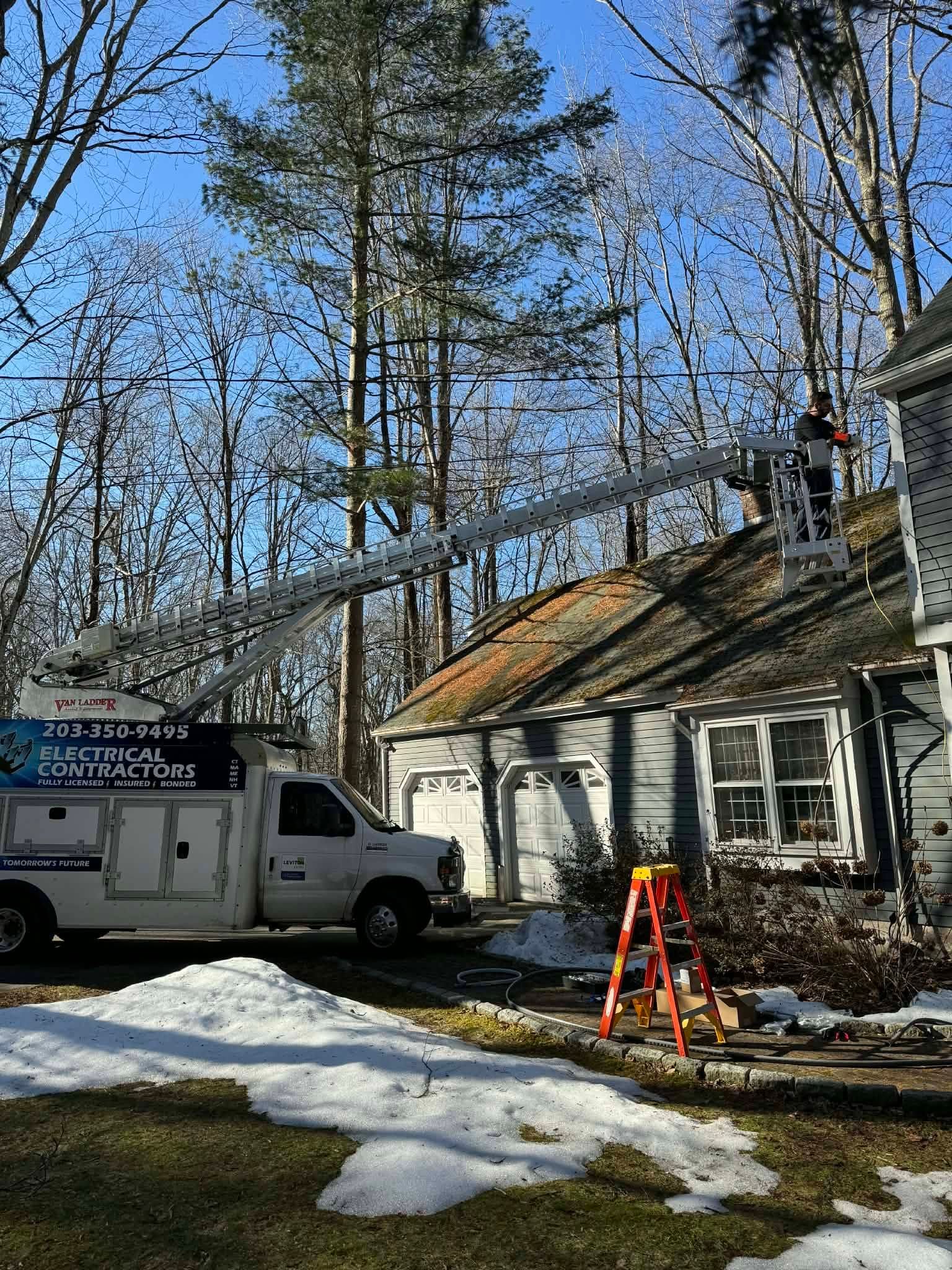 Worker on a ladder near a house roof with snow patches, a white truck, and tall trees in the background