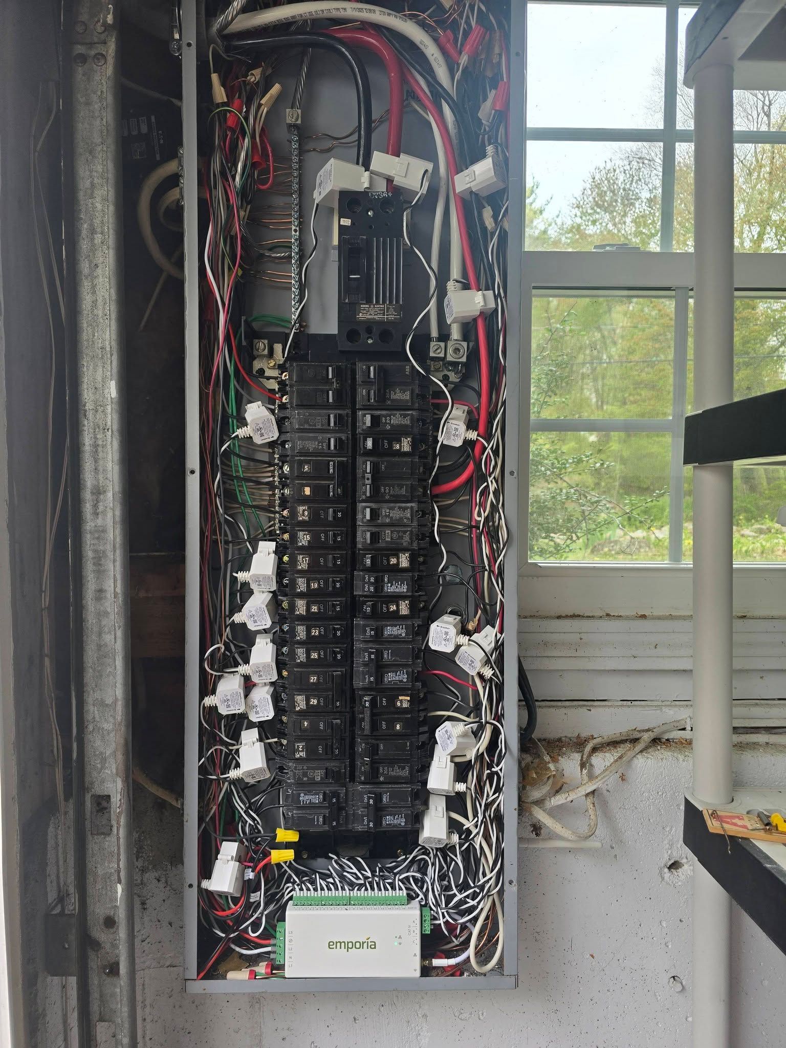 Open electrical breaker panel with exposed wiring beside a window in a damaged wall