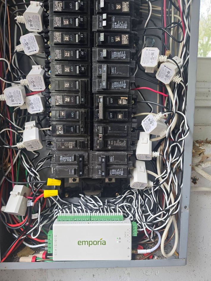 Open electrical panel with rows of black breakers and tangled white, red, and black wiring.