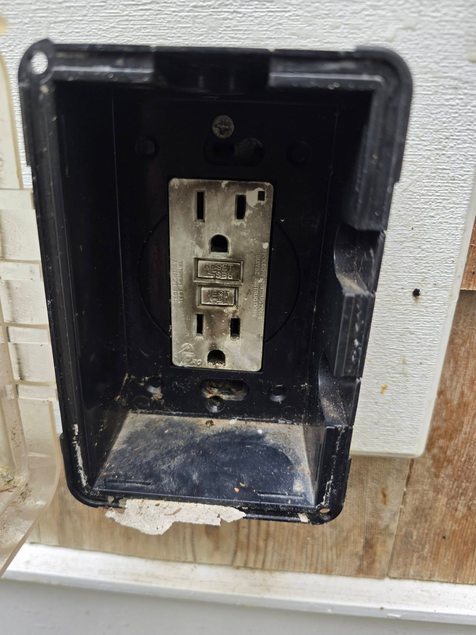 Black recessed outdoor outlet box with a weathered duplex receptacle on a wooden wall.
