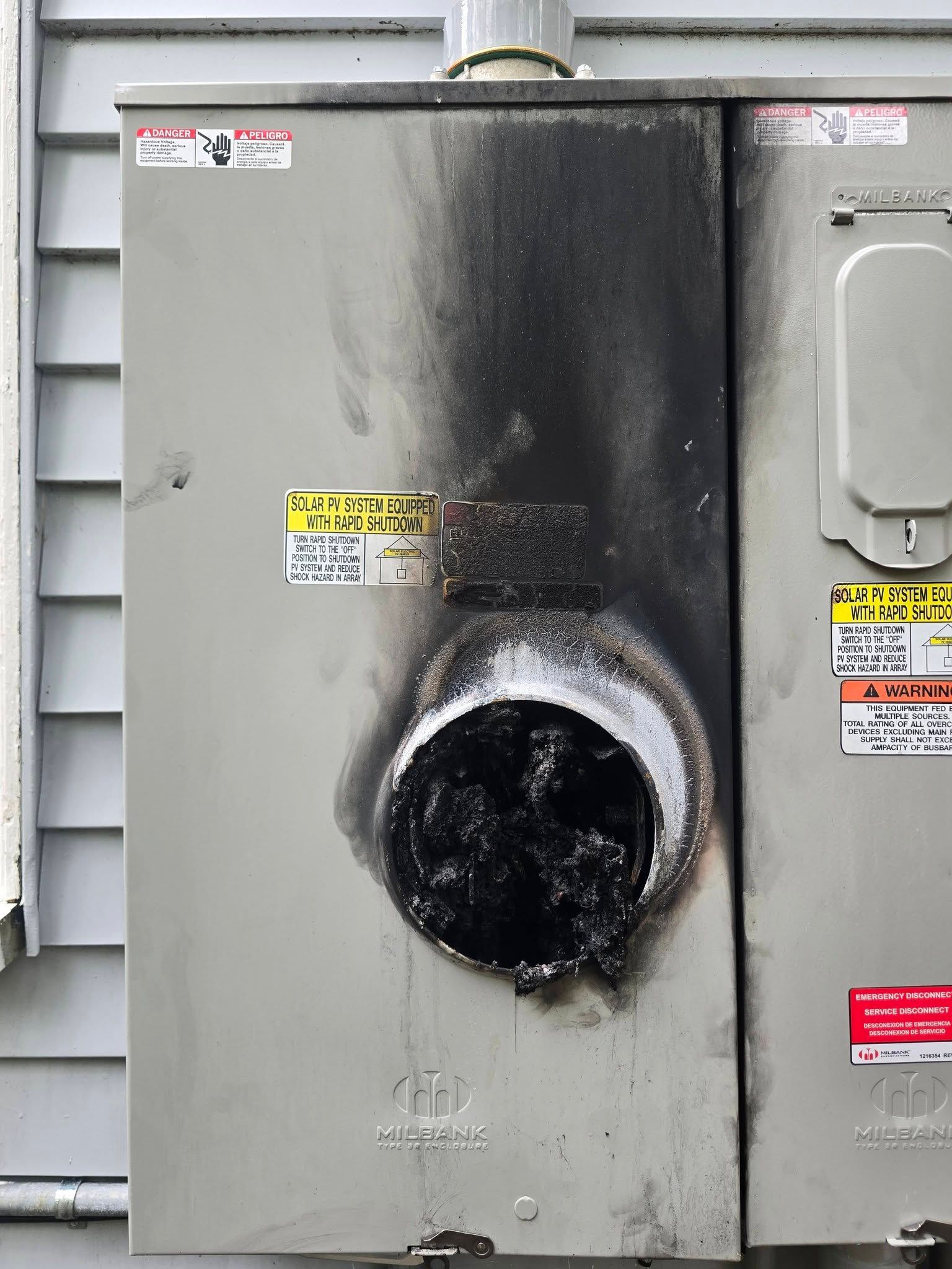 Gray electrical panel with a burned, blackened circular vent or pipe opening in the center.