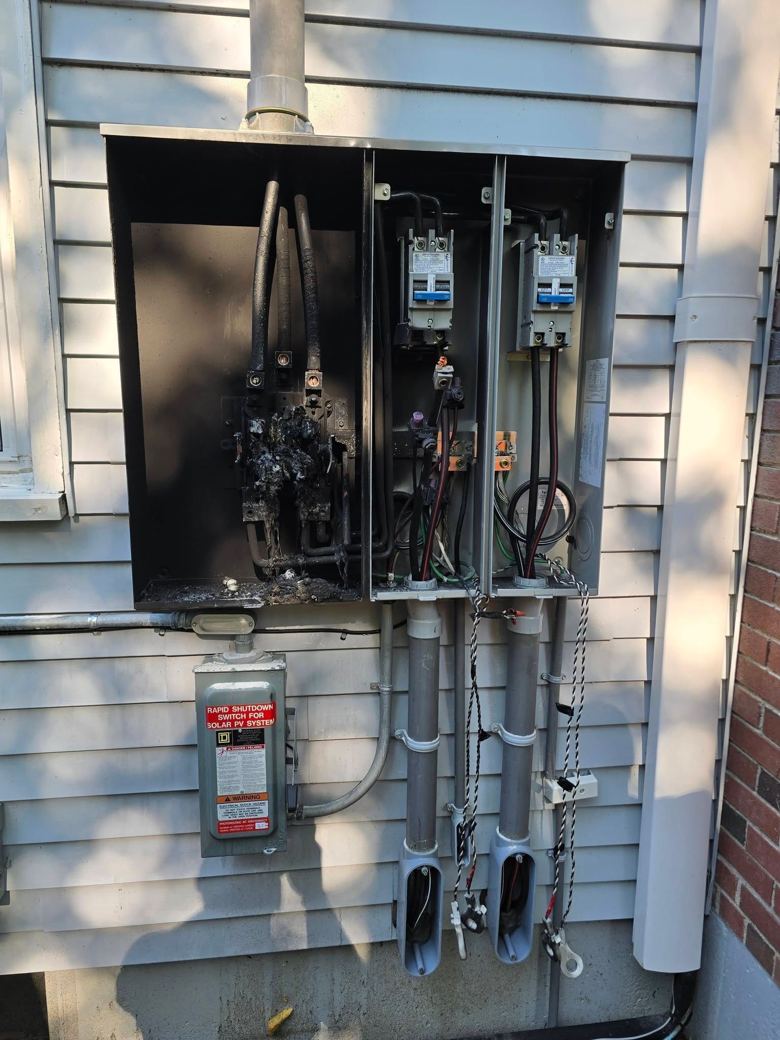 Open electrical meter panel on house exterior with charred wiring and smoke damage