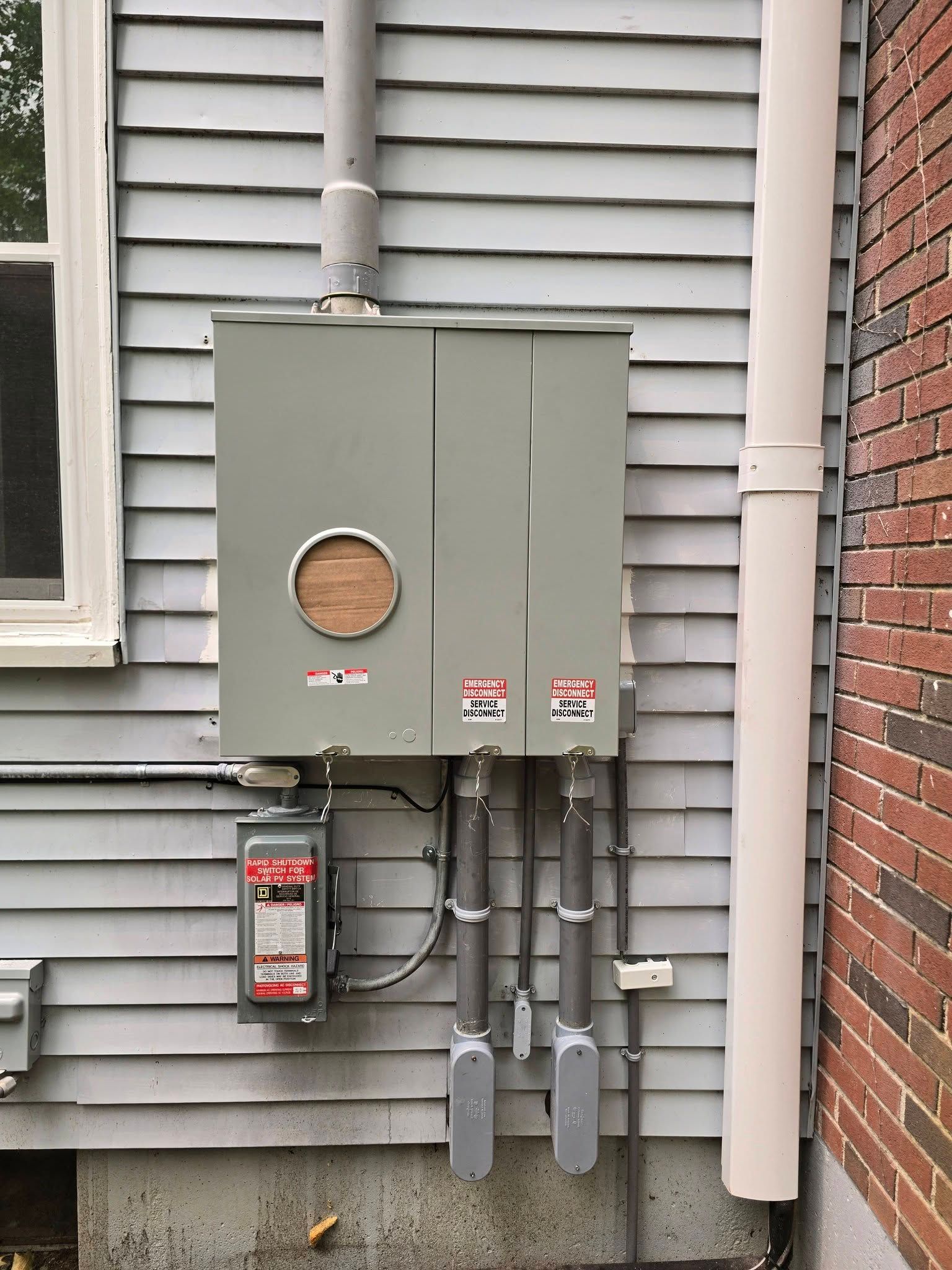 Outdoor utility meter and electrical service boxes mounted on a house wall with gray siding and brick trim
