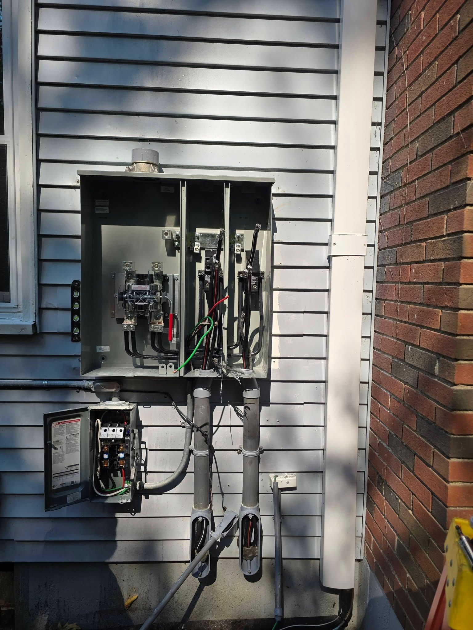 Open electrical panel and meter box mounted on a white house exterior beside a brick wall.