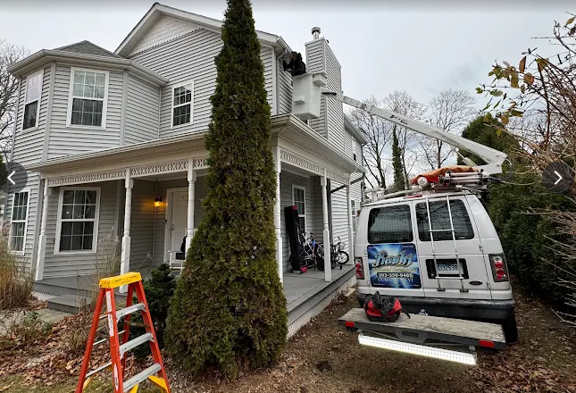 Workers and equipment beside a gray house with a ladder and utility van in the yard