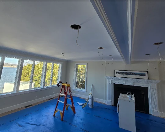 Empty living room under renovation with blue floor covering, ladder, large windows, and fireplace