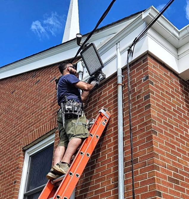 Worker on ladder installing a floodlight on a brick house exterior