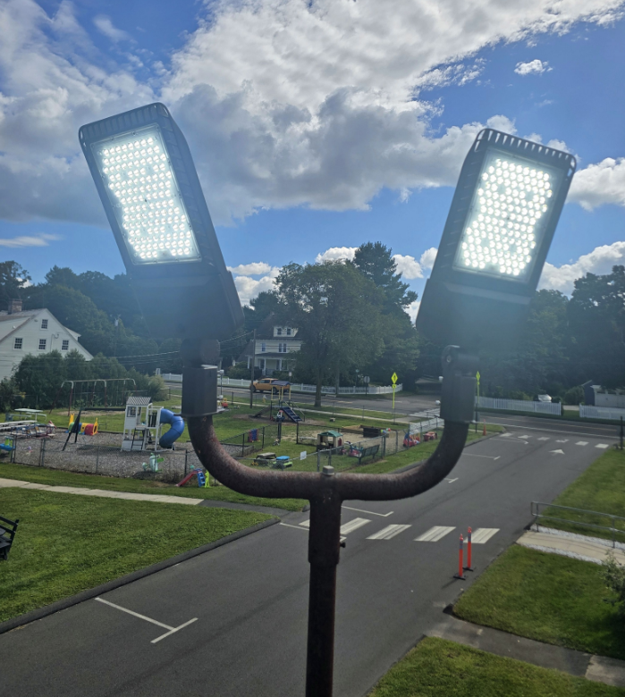 Two bright LED floodlights mounted on a pole, overlooking a park and road under a cloudy sky.