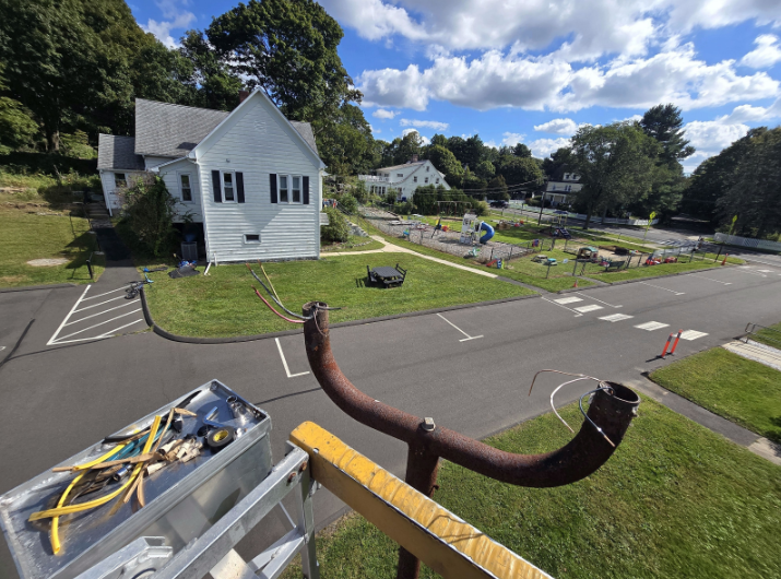 White church beside a road and cemetery, seen from a rooftop with a large rusted pipe in the foreground