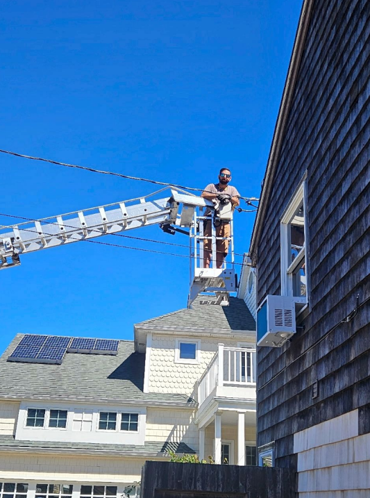 Firefighters on a ladder truck work on a shingled house roof under a clear blue sky.