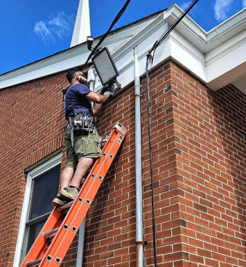 Technician on an orange ladder installing a floodlight on a brick house exterior