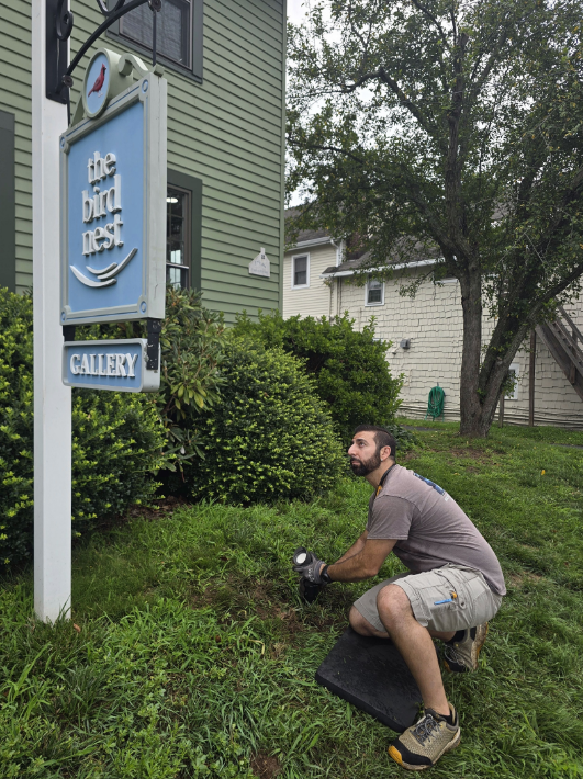 Man crouches by a blue sign in front of a green house and shrubs.