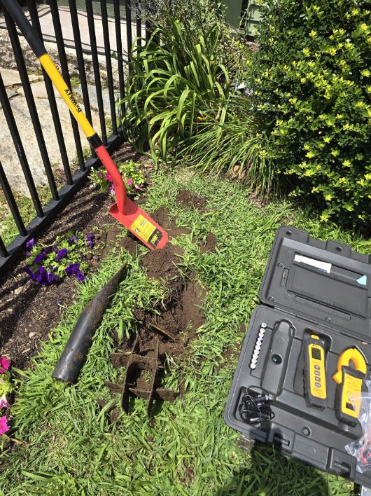Backyard garden tools and a dug-up hole beside a black fence, with a toolbox and spade nearby