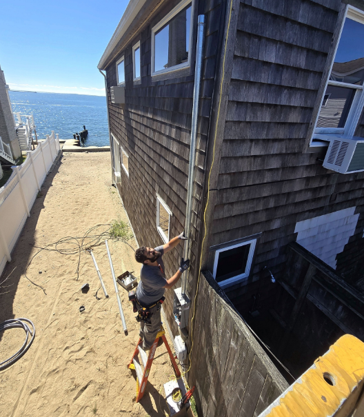 Person on a ladder working beside a shingled house with ocean view nearby