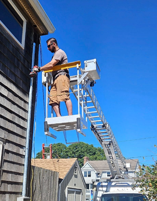 Worker on an elevated platform beside a house, using a ladder lift under a bright blue sky.