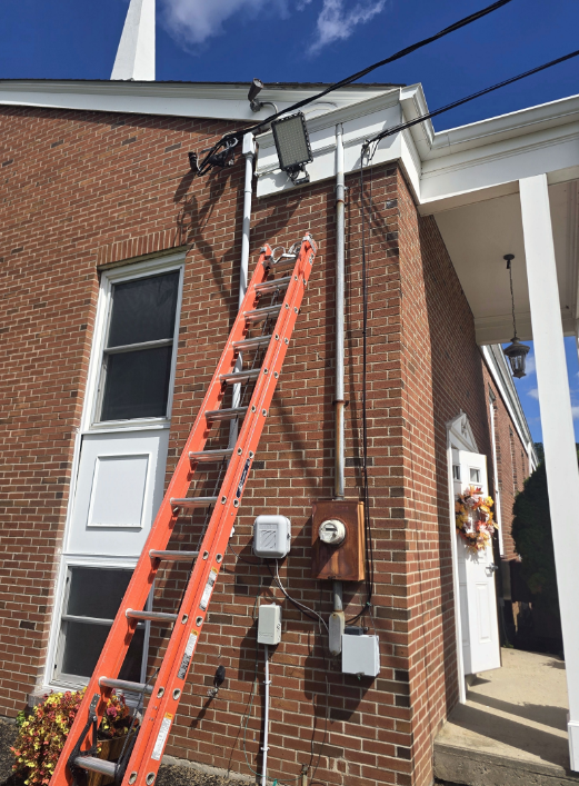 Ladder leaning against brick building below utility lines, with electrical meters and an alleyway entrance