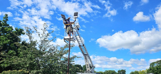 Crane lifting a worker high above green trees under a bright blue sky with clouds