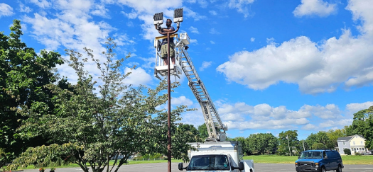 Utility worker on a lift near a tall pole by a road, under a blue sky with clouds.