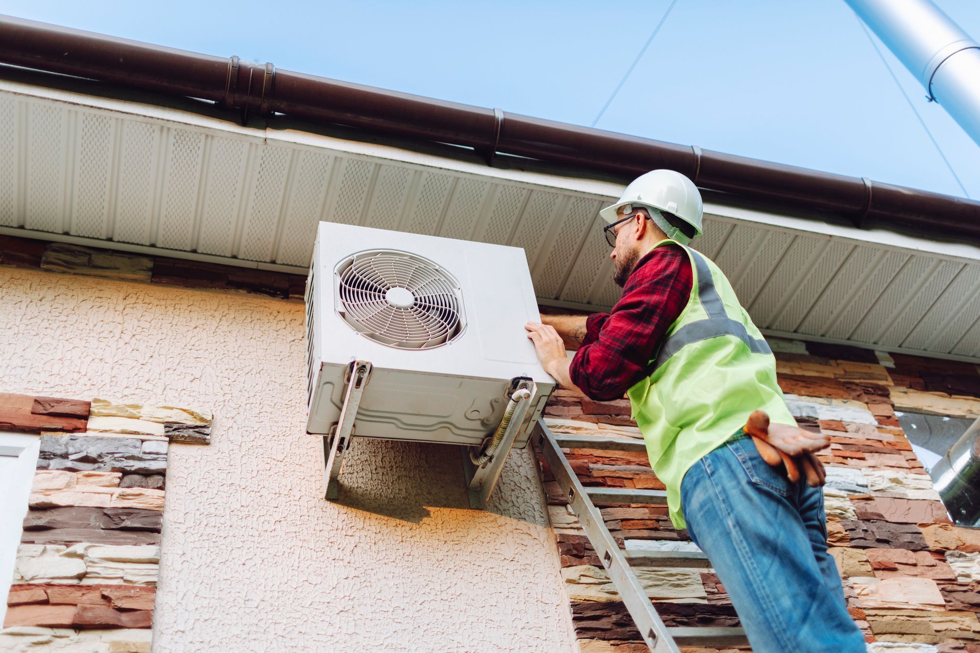 HVAC technician in hard hat and vest on a ladder repairing an outdoor air conditioning unit.