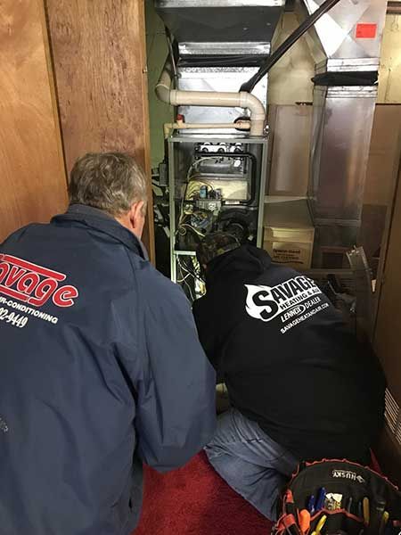 Two technicians in Savage jackets work on a furnace in a basement.
