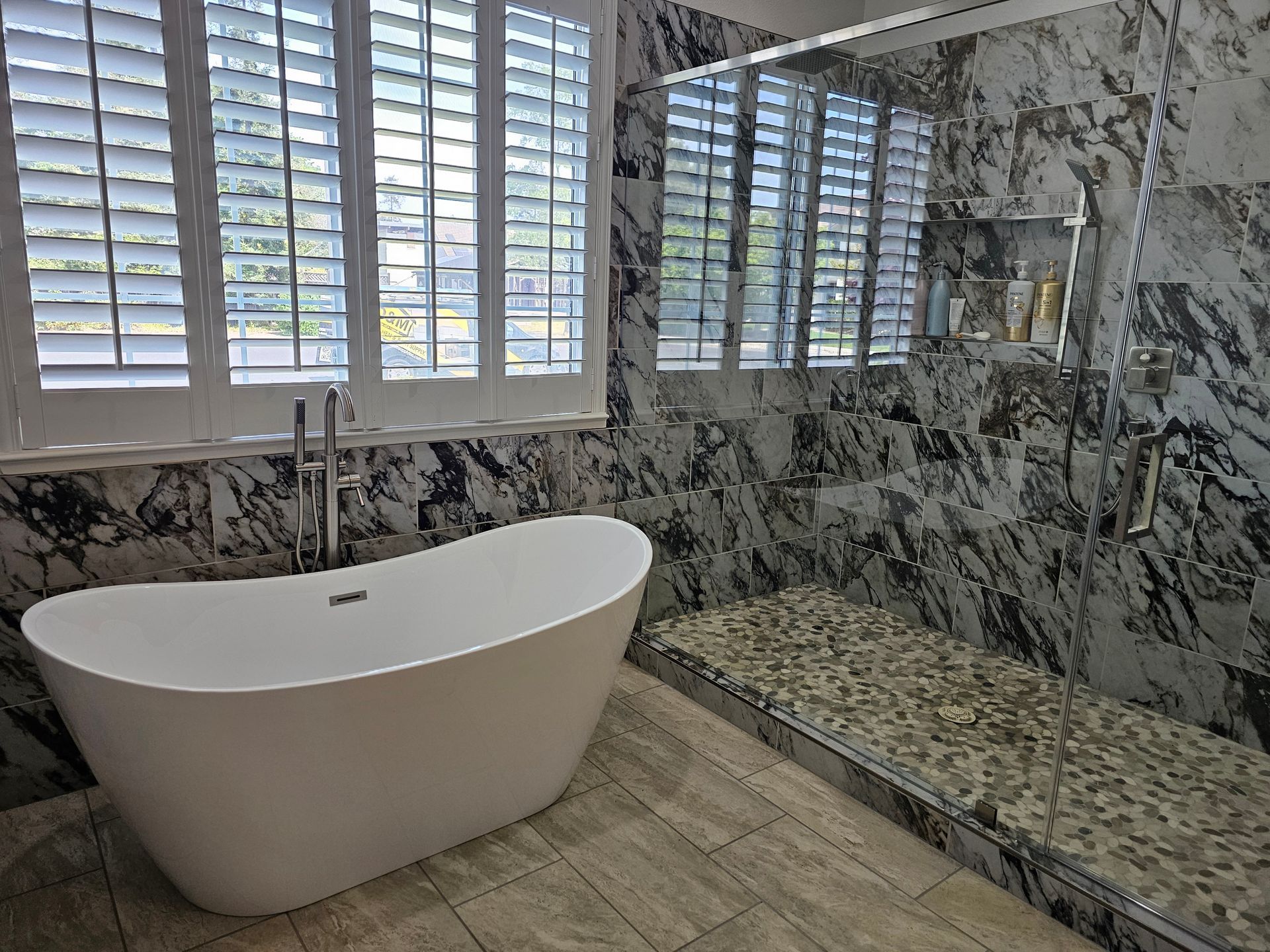 Elegant bathroom with freestanding white tub, marble walls, and window shutters overlooking a large shower bench