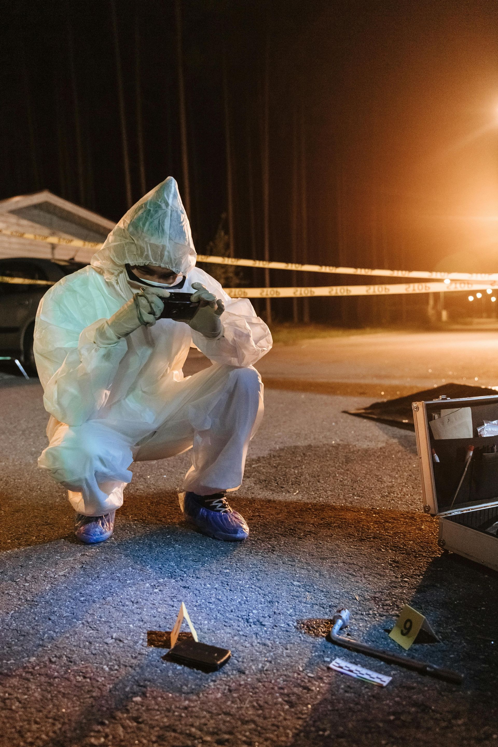 A forensic investigator in a white protective suit crouches to photograph evidence at a cordoned-off crime scene at night.