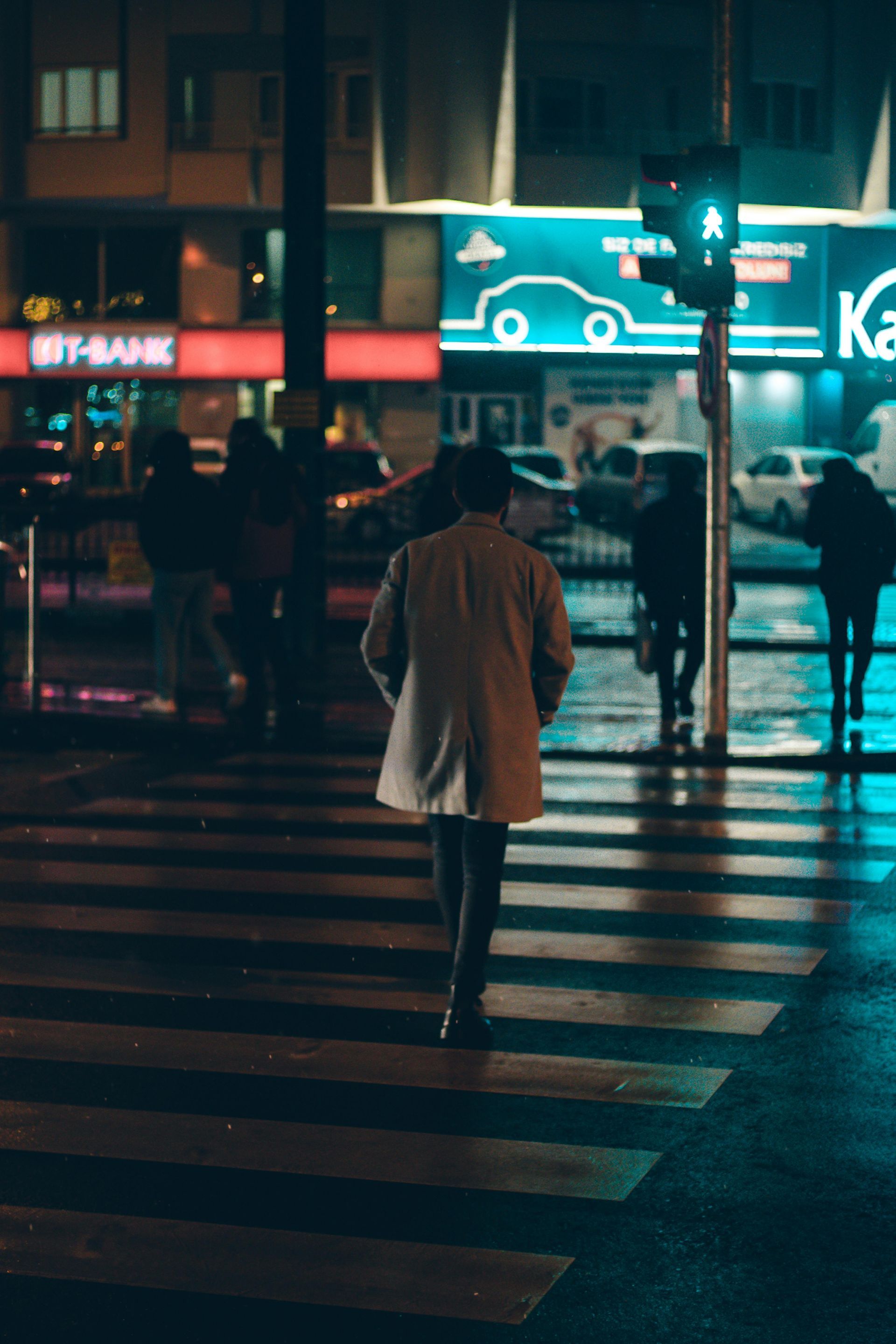 A person in a light coat walks across a wet city crosswalk at night near a glowing green pedestrian signal.