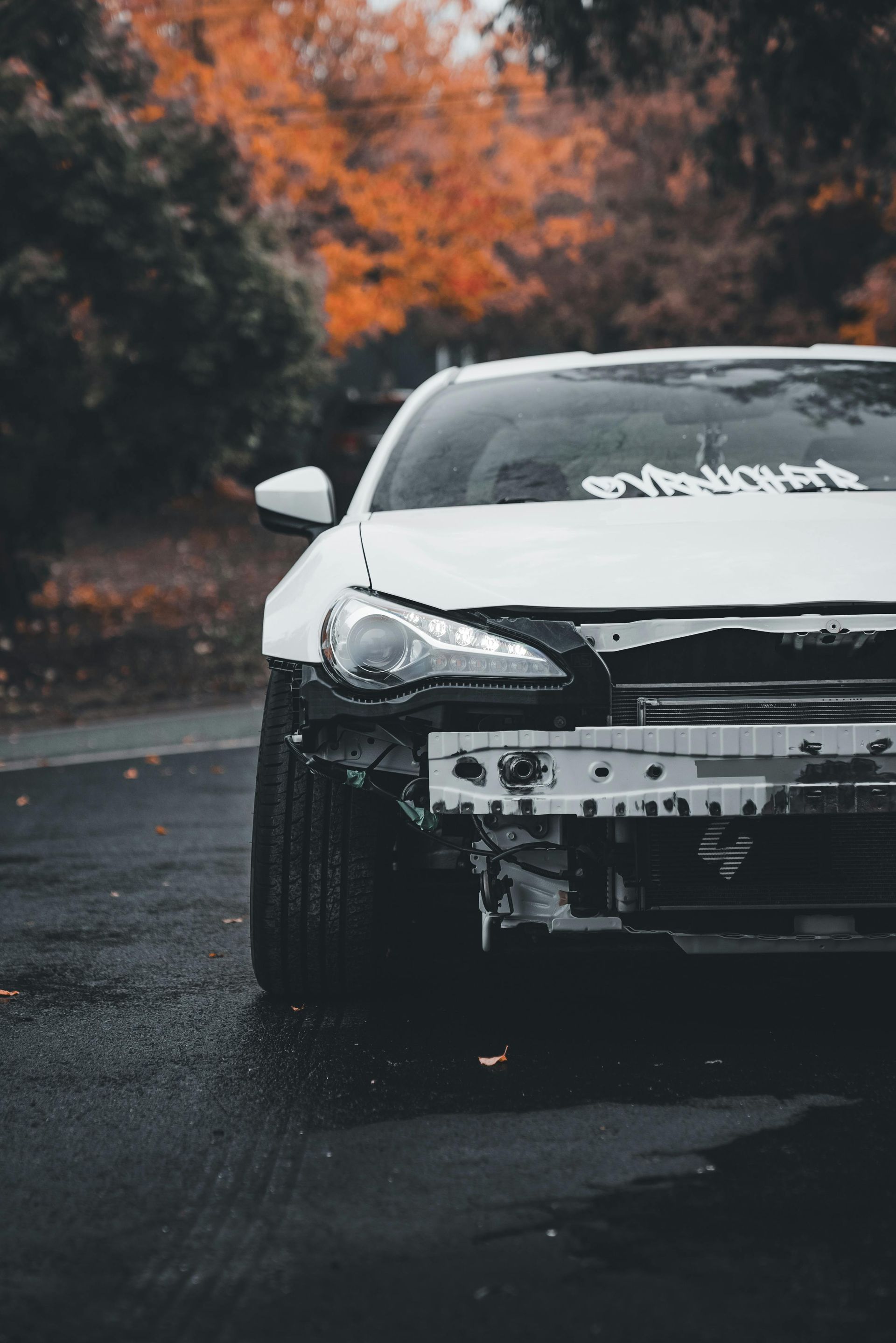 A white car with a missing front bumper sits on a wet road, framed by autumn trees in the background.