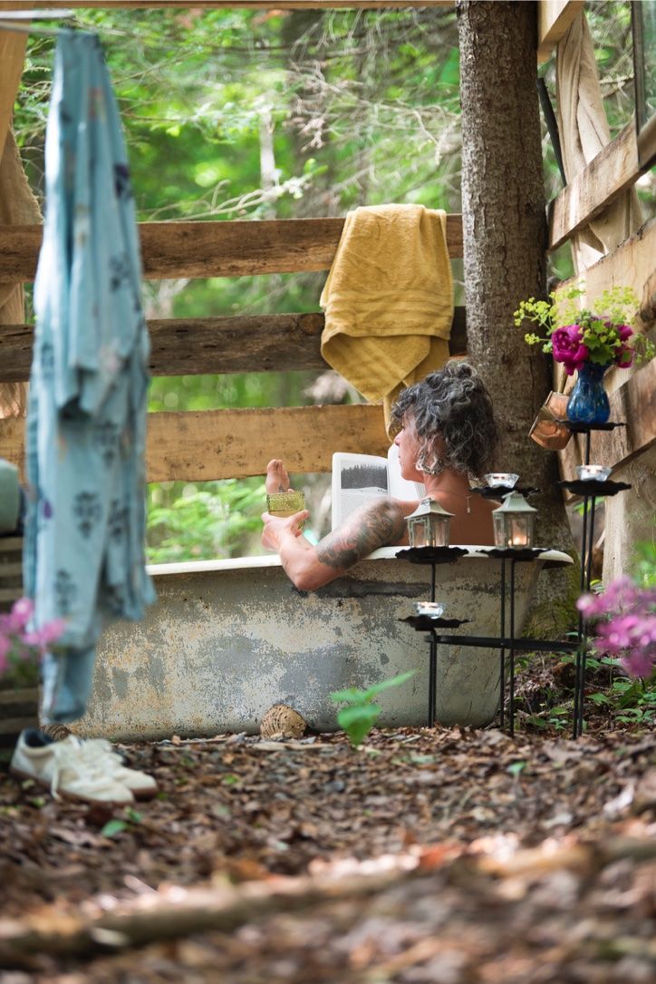 Person in a bathtub outdoors, surrounded by nature and candles, with a robe and towel nearby.