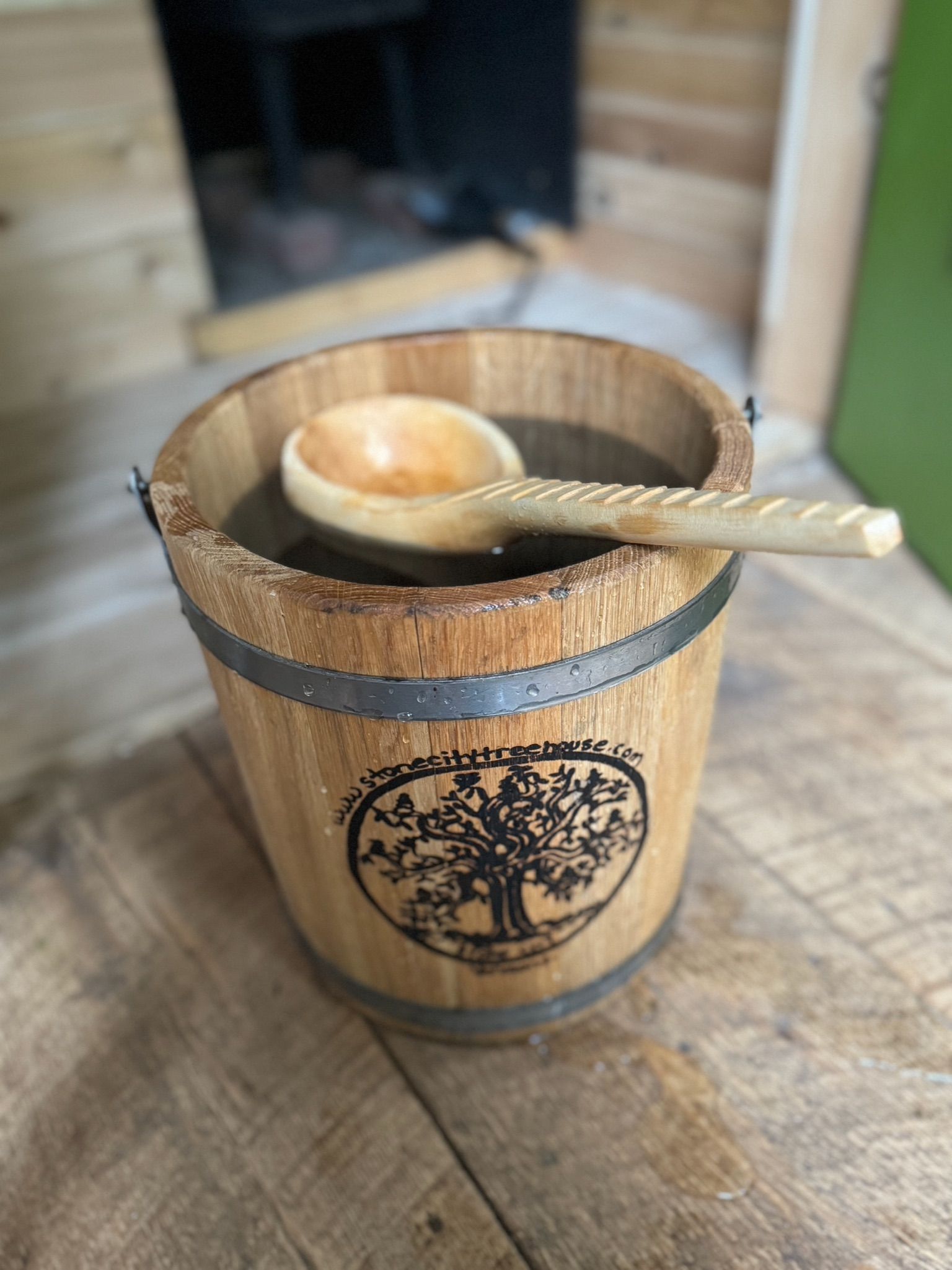 Wooden bucket with a wooden spoon resting inside, sitting on a wooden surface, with a blurred fireplace in the background.
